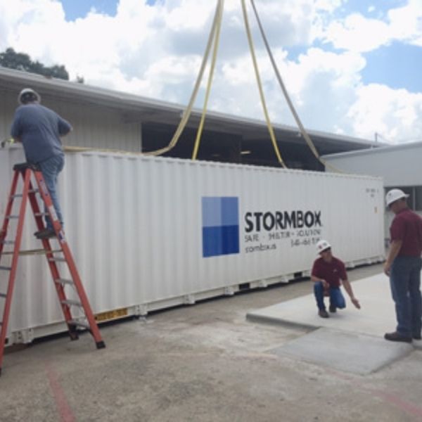 A large modular storm shelter being lowered by a crane onto a concrete foundation.